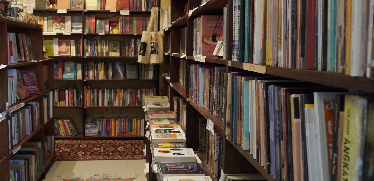 Narrow bookstore aisle lined with shelves of books on both sides, with stacks of books on the floor.