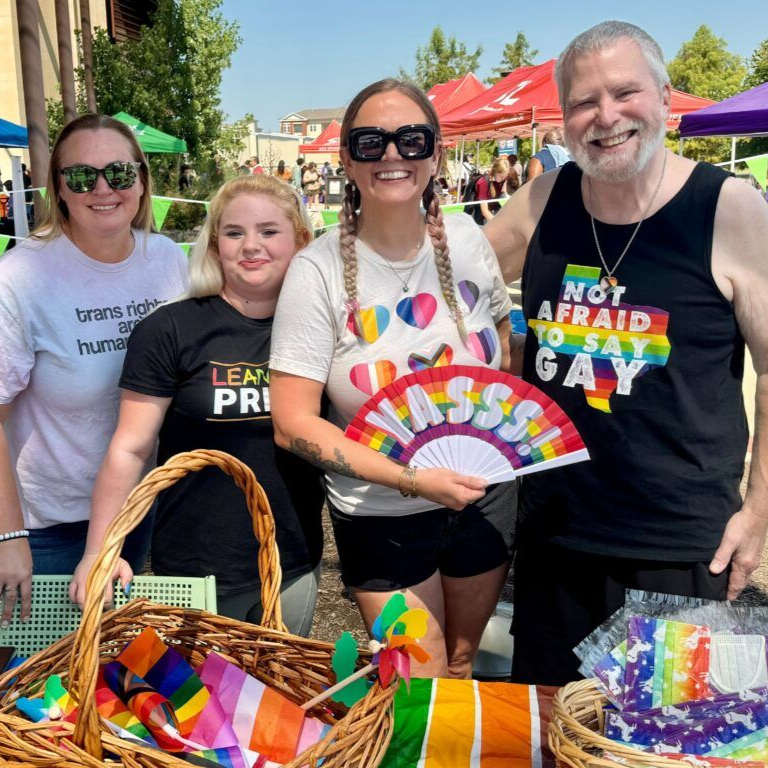 Four people pose smiling at an outdoor pride event, holding a 