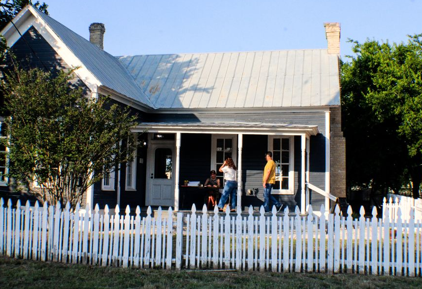 A dark blue, single-story house with a white picket fence, metal roof, and three people on the porch.