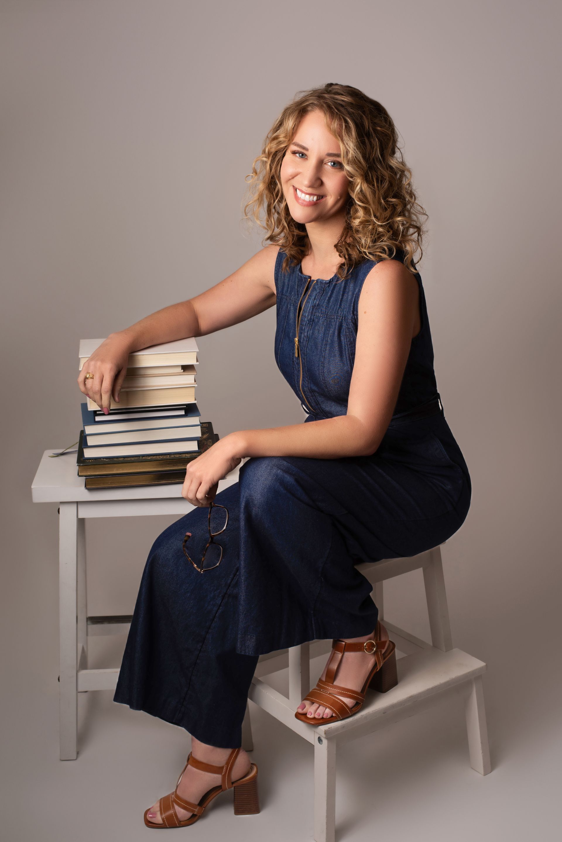 Woman in denim jumpsuit sits on a stool, smiling, with a stack of books.