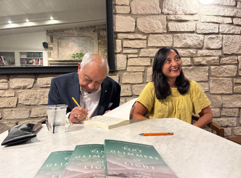 Man signing a book; woman smiles. Three books on a table, brick wall background.