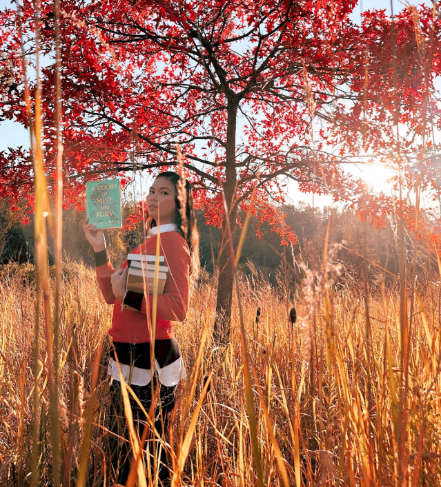 Person standing in tall grass, holding books, with vibrant red tree in background.