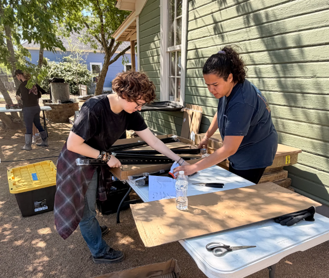 Two people work at a table outdoors beside a house, handling papers and materials in the shade.