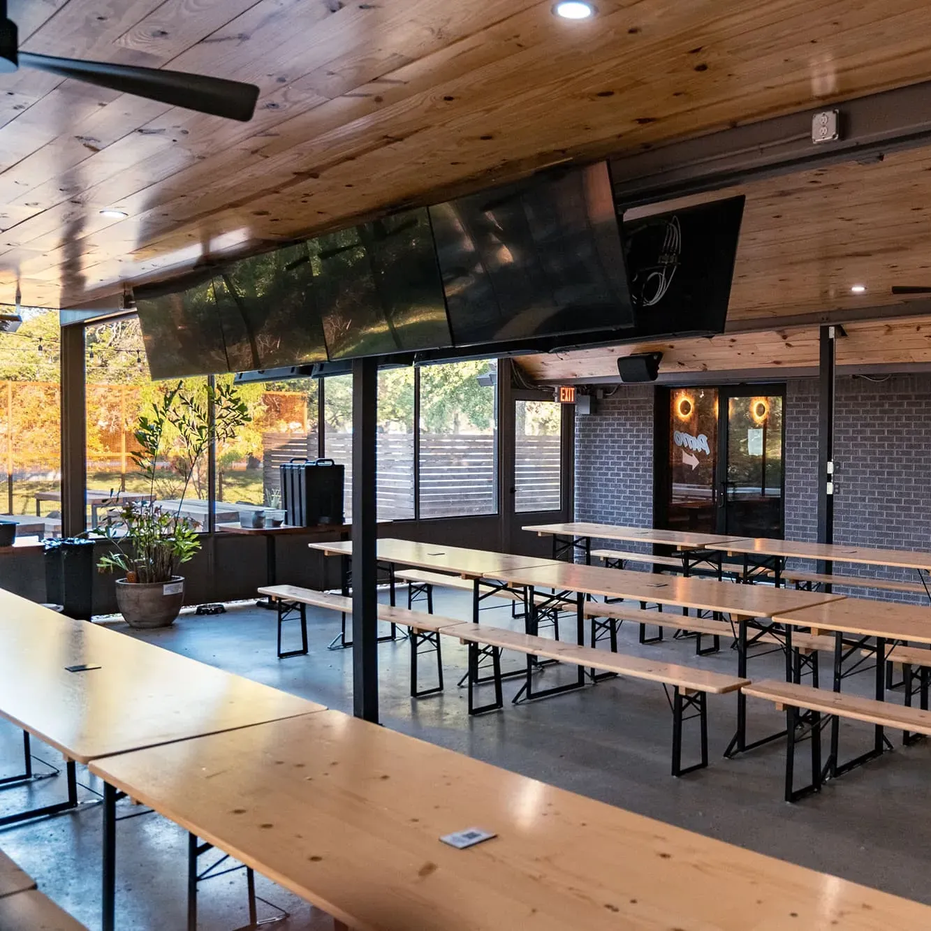 Indoor-outdoor bar space with long wooden tables, many TVs, and a large window overlooking trees.