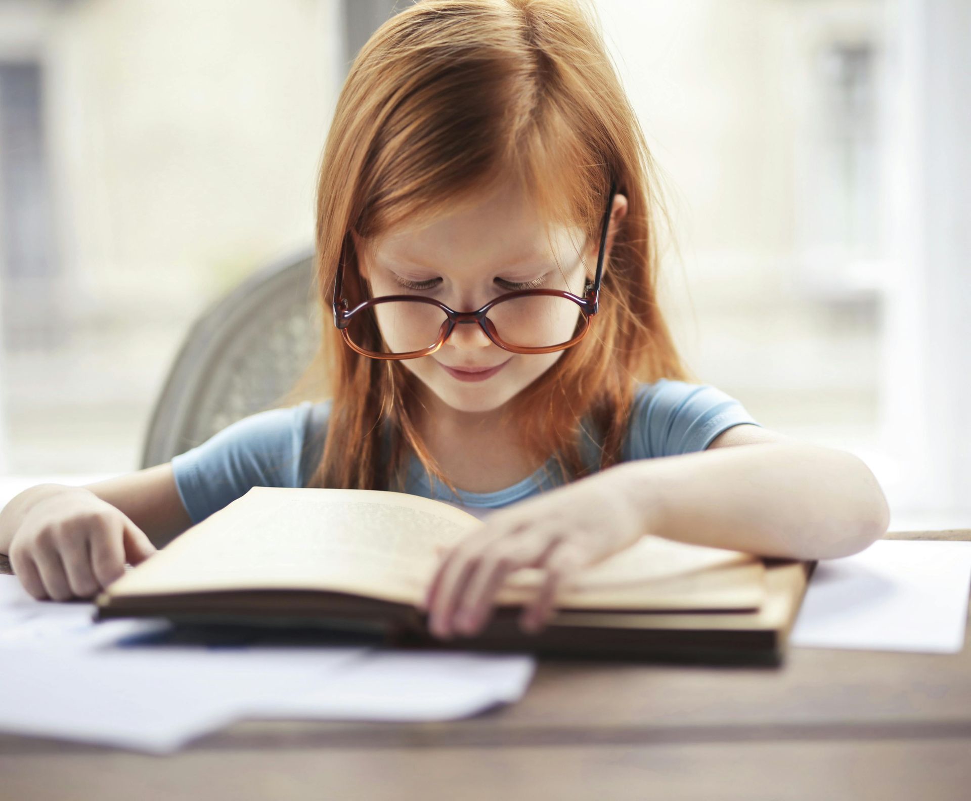 Child reading a book at a table, wearing glasses in a bright room