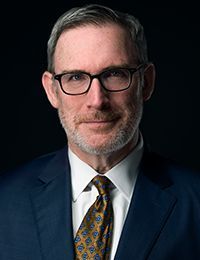 Man wearing glasses and a suit, looking at the camera with a neutral expression. Dark background.