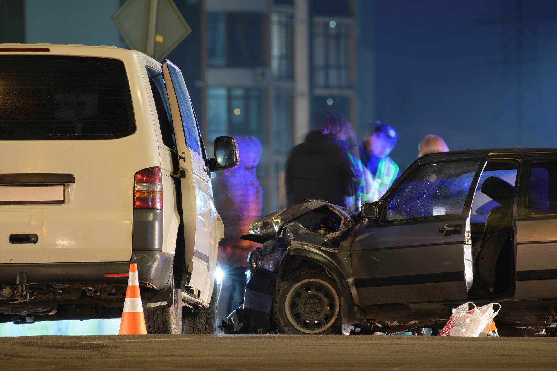 Car accident scene at night; damaged vehicles, emergency responders, and a traffic cone.