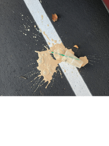 Spilled tan liquid with straw and debris on black asphalt with white line.