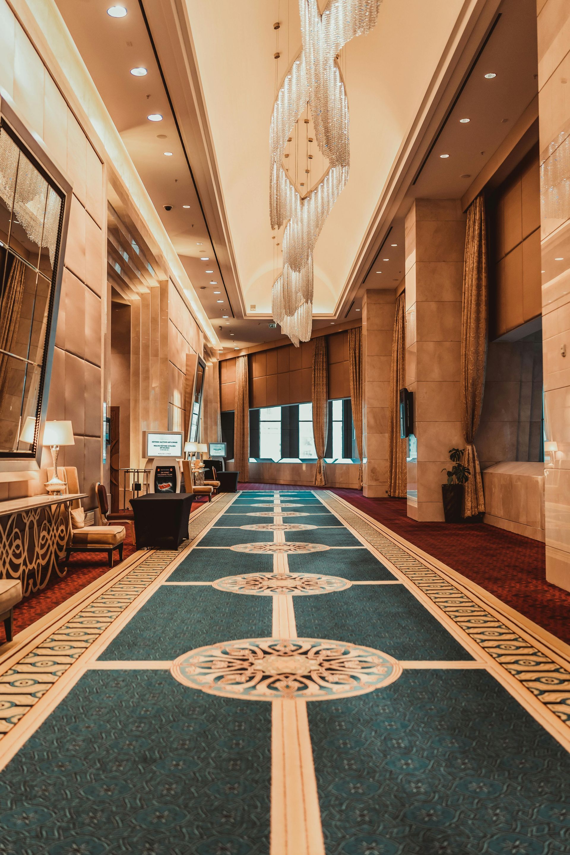 Long hotel hallway with patterned blue carpet, columns, and chandelier.
