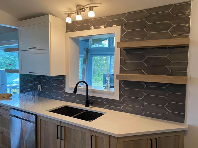 Un évier de cuisine avec un robinet noir, des comptoirs blancs, une crédence en carreaux hexagonaux gris et des étagères flottantes en bois clair.