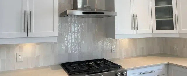 A modern kitchen featuring white cabinets, a gas stove, stainless steel range hood, and glossy, light-colored tile backsplash.