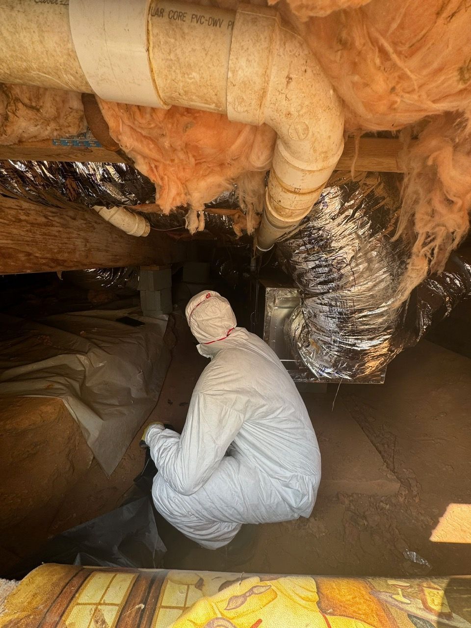 A man in a white suit is kneeling down in an attic looking at a pipe.