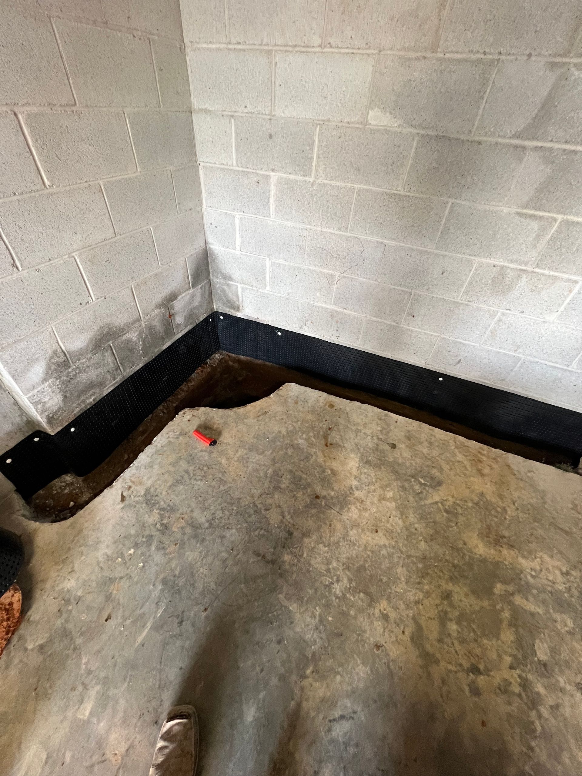 Basement corner with black drainage channel along the wall, concrete walls, dampness visible.