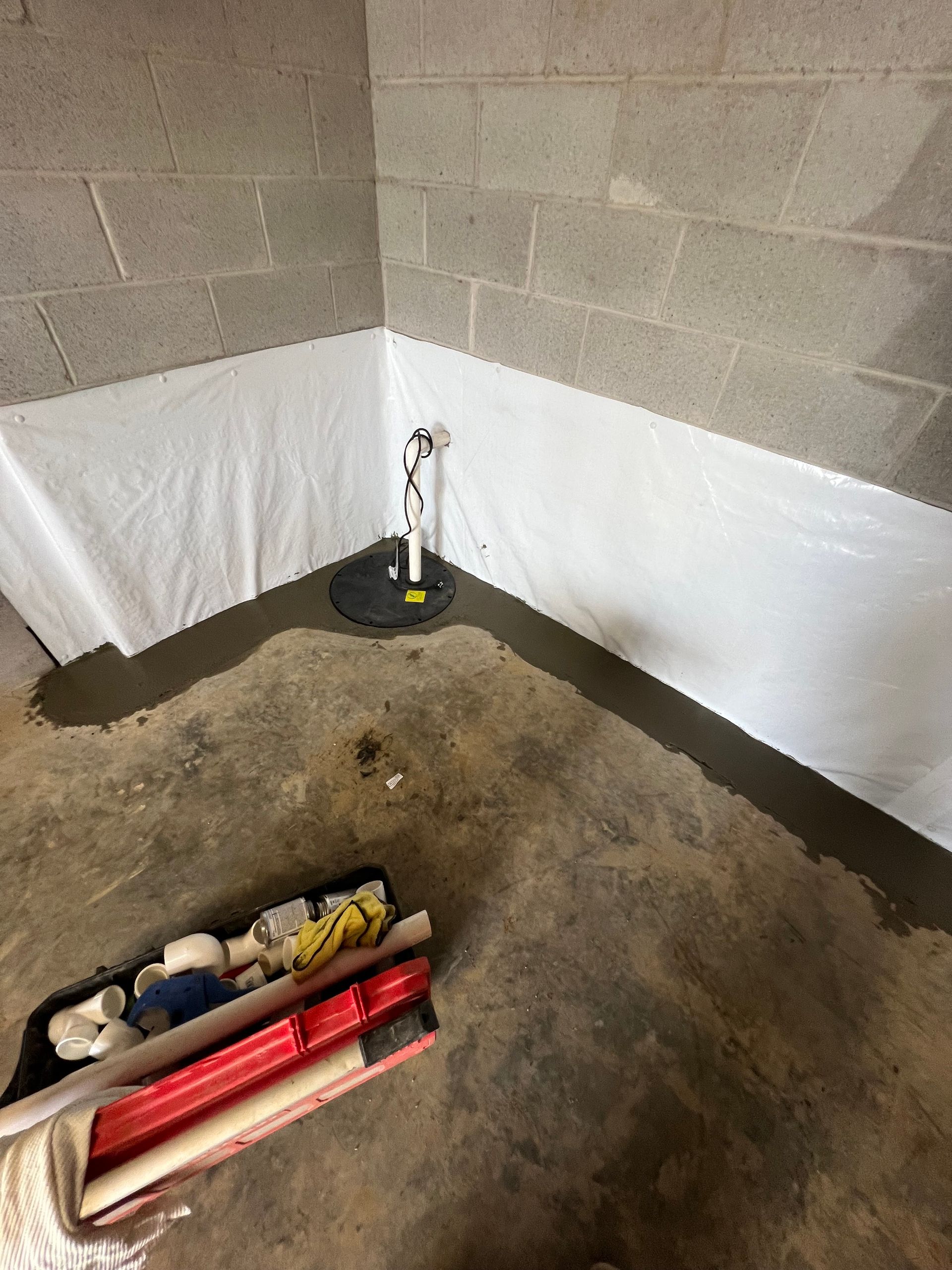Basement corner with a sump pump and white waterproof lining on the walls. Tools lie on the concrete floor.