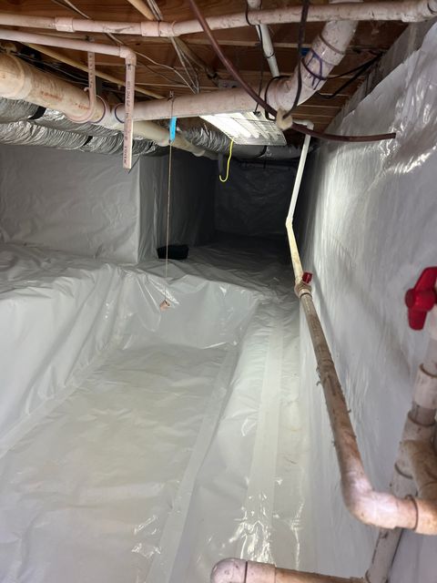 Crawl space with white vapor barrier. Pipes and wires are visible, with dim lighting.