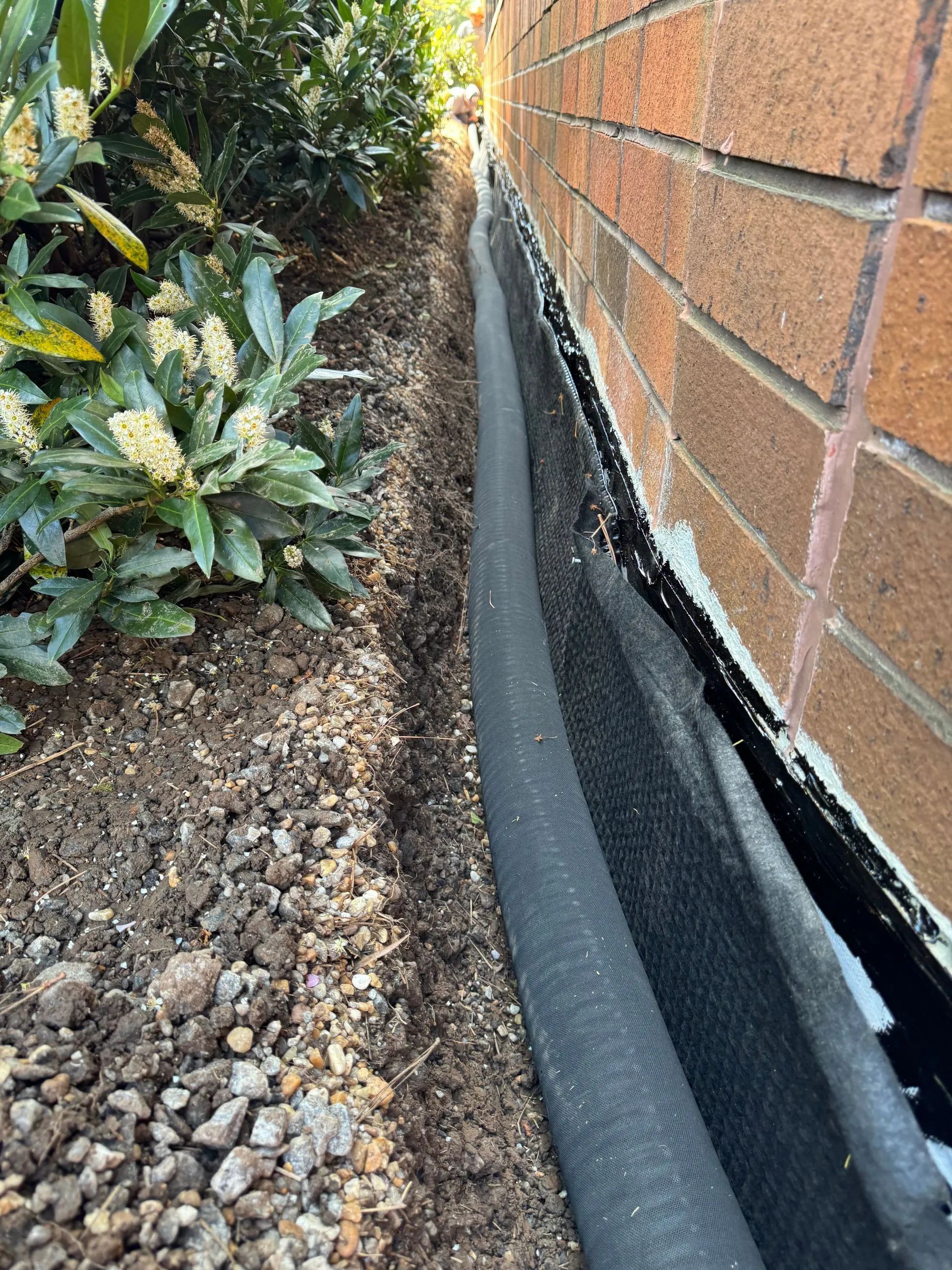 Gravel trench next to a brick wall with black fabric lining. Green shrubbery to the left.