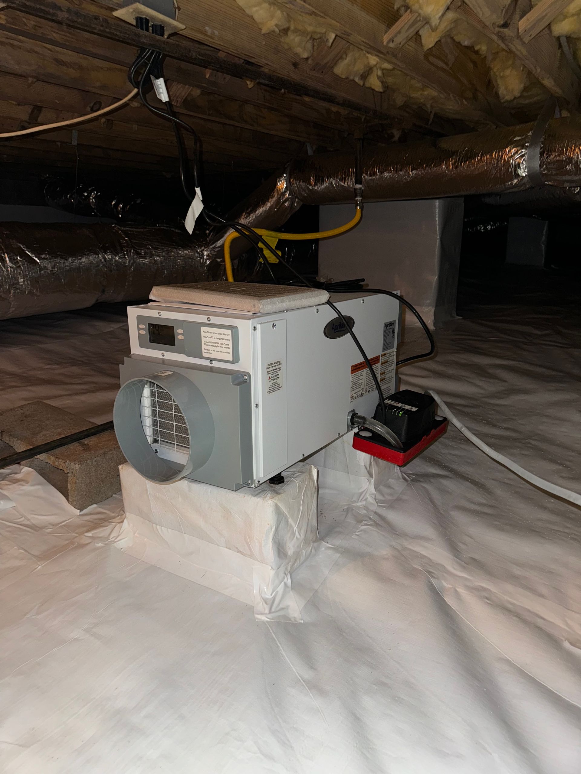 Dehumidifier in a crawl space, mounted on cinder blocks, surrounded by white plastic sheeting.