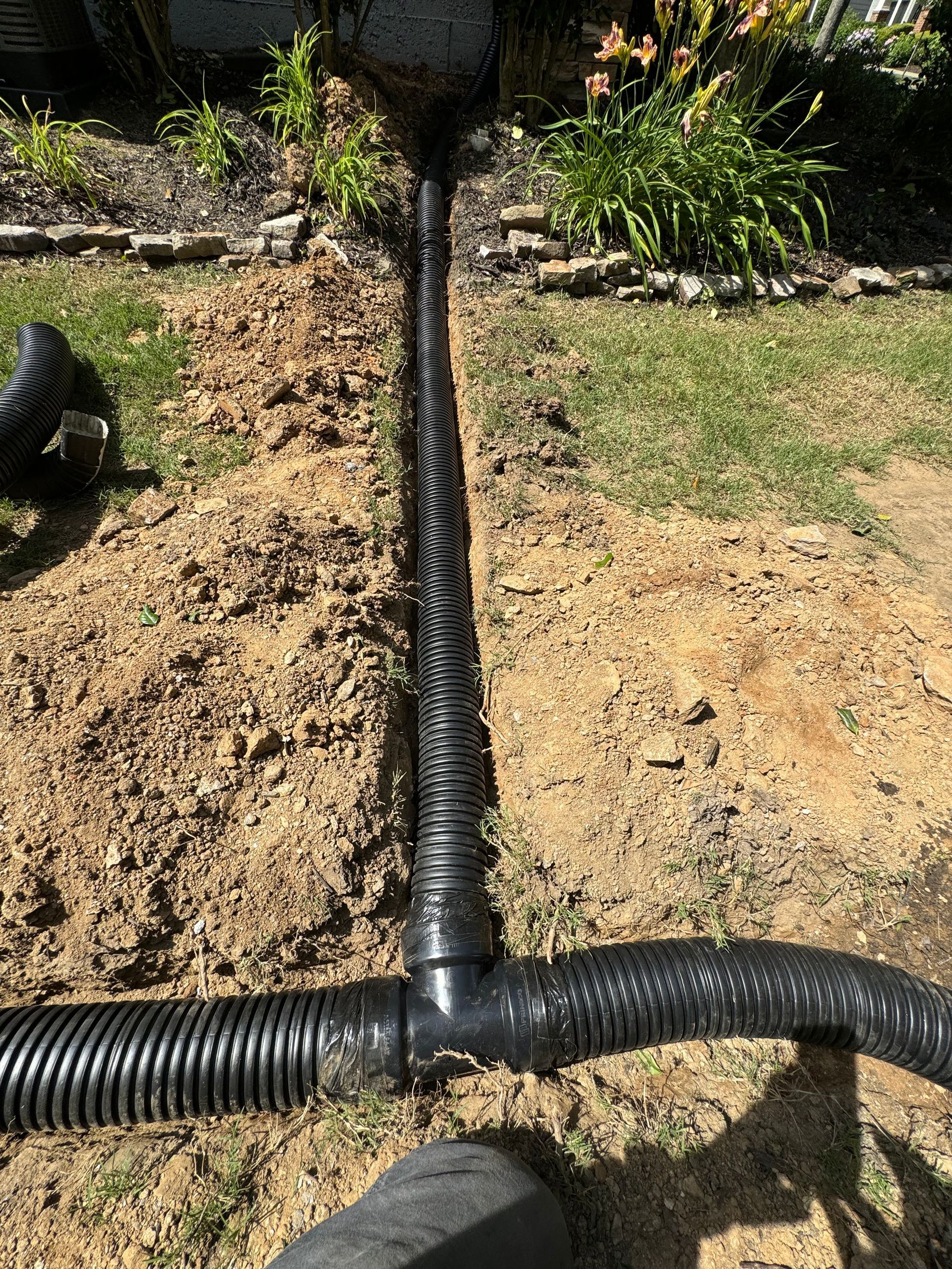 Black corrugated drain pipes installed in a trench in a yard, connecting to a downspout and branching off.