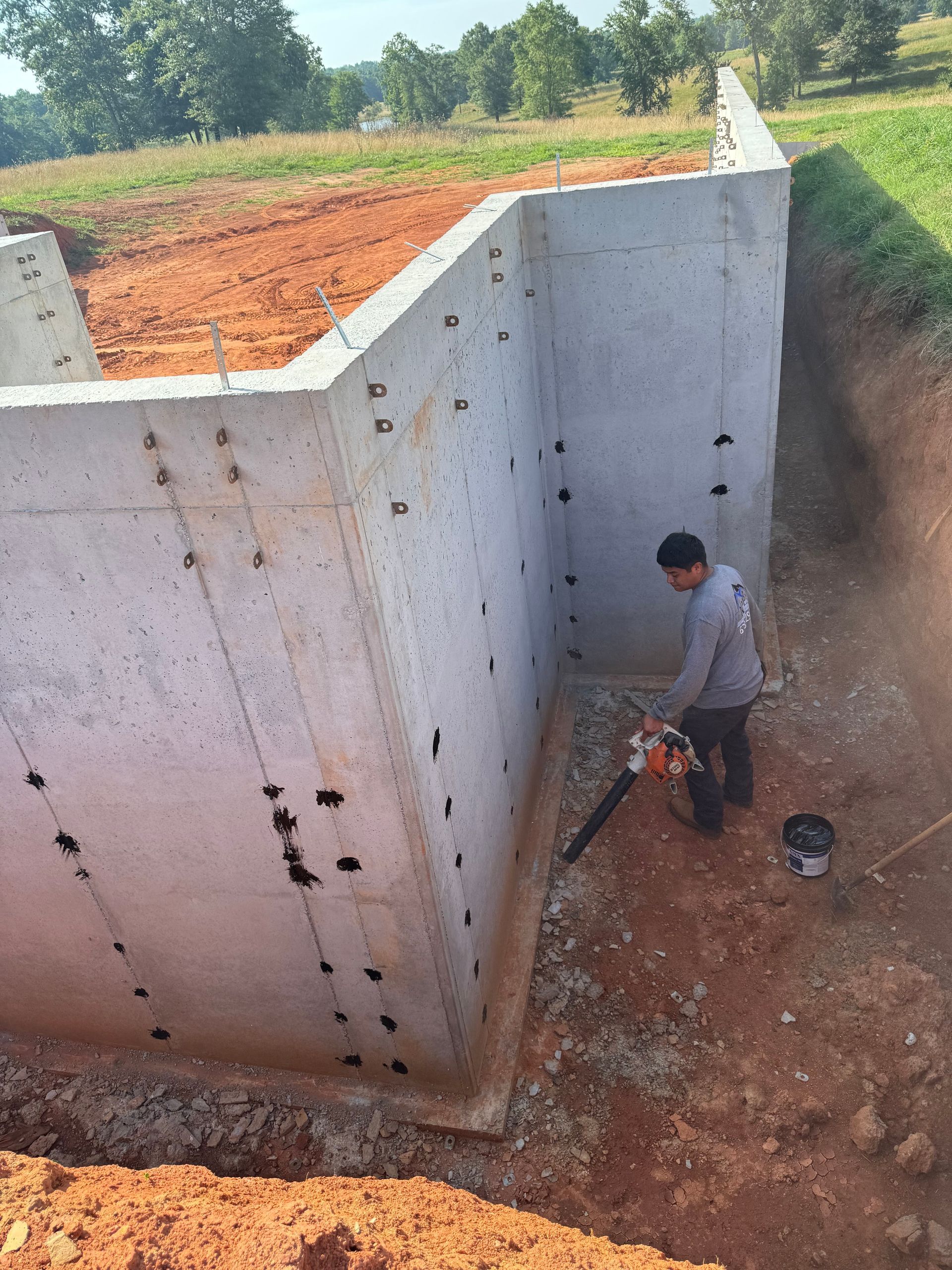 Man using a saw to cut concrete foundation wall. Outdoors, brown earth and green grass.