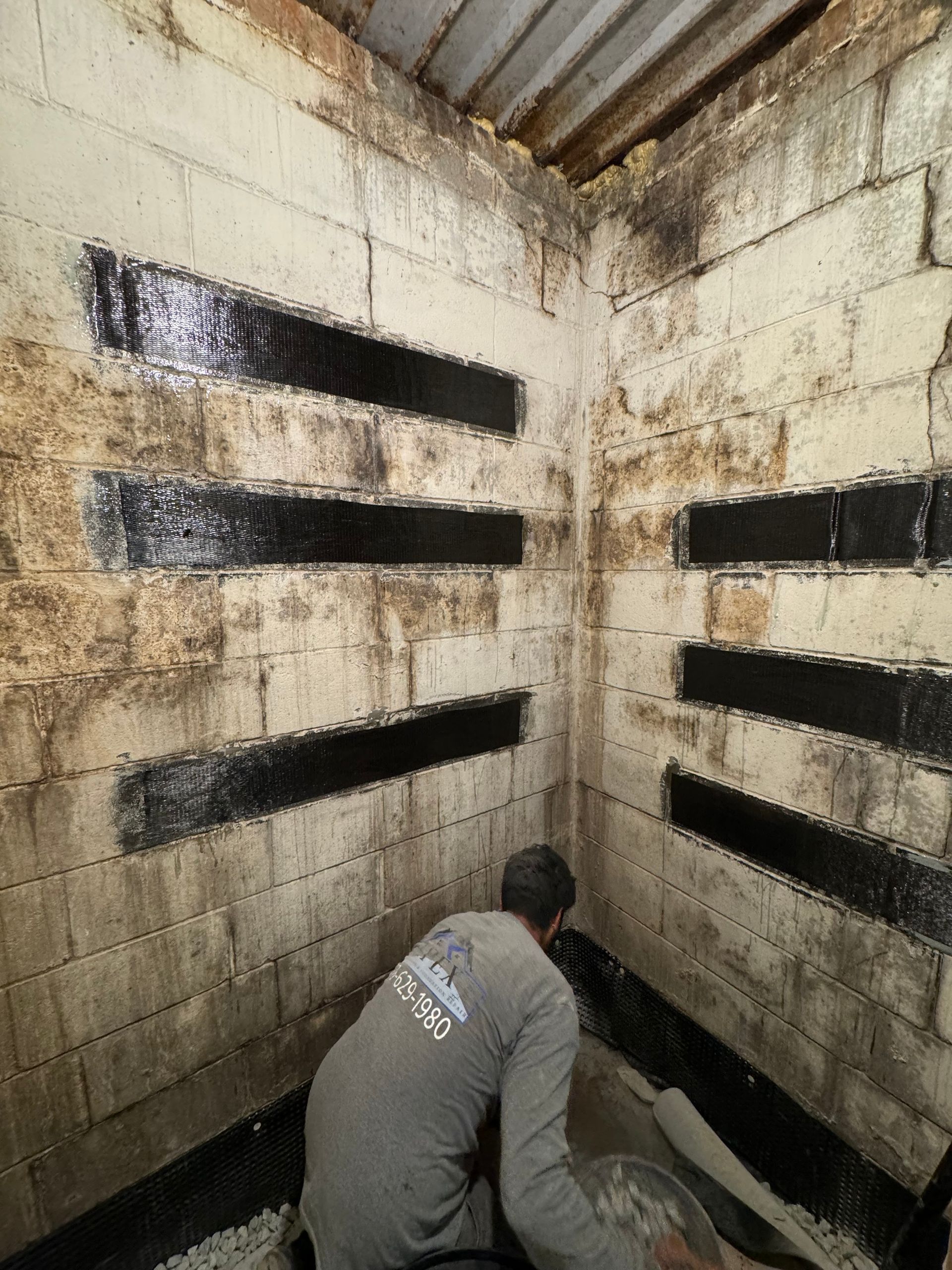 Man installing black strips on cinder block walls in a damp basement corner.