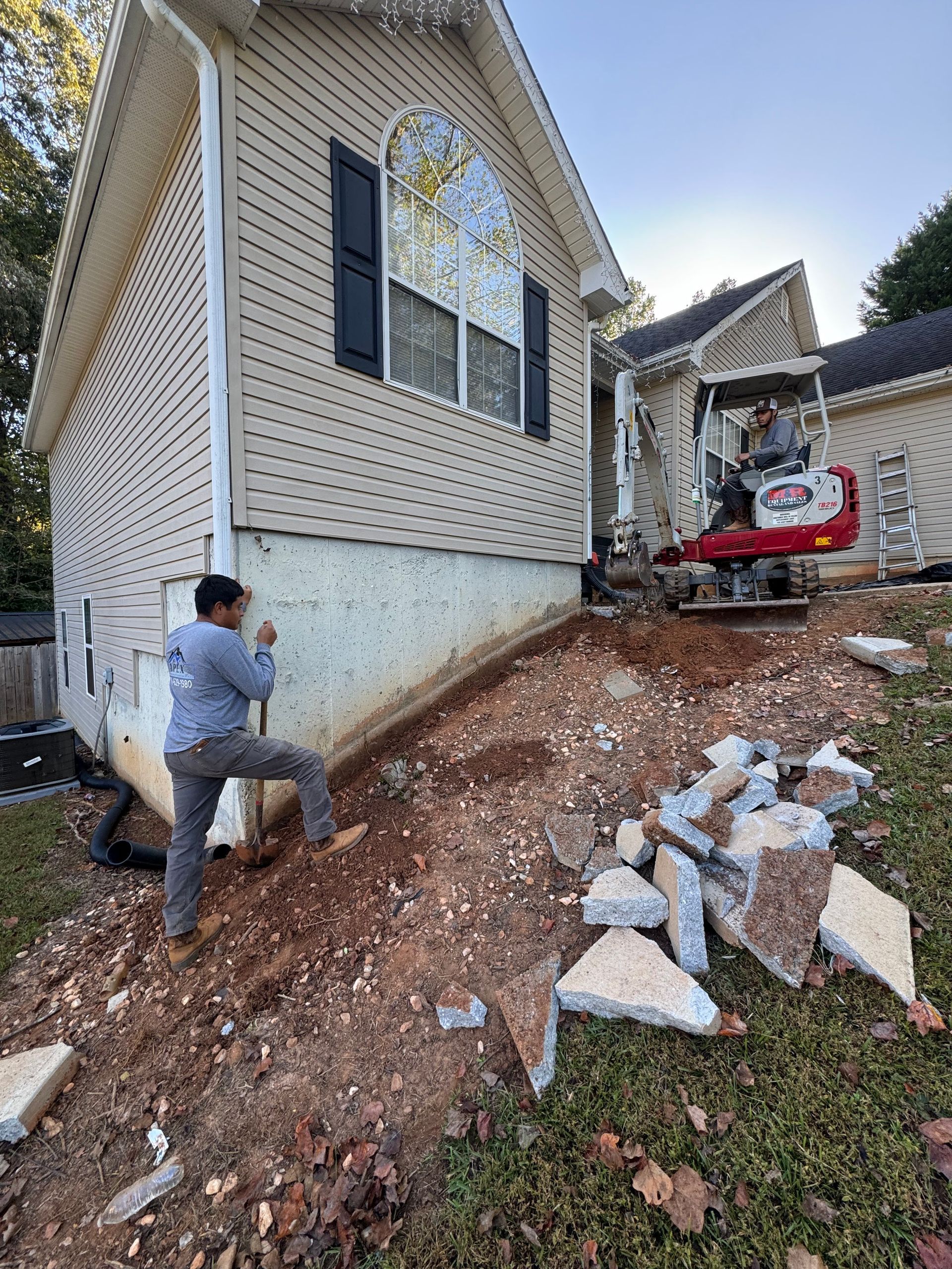 Construction workers excavating dirt near a house foundation; one uses a tool.