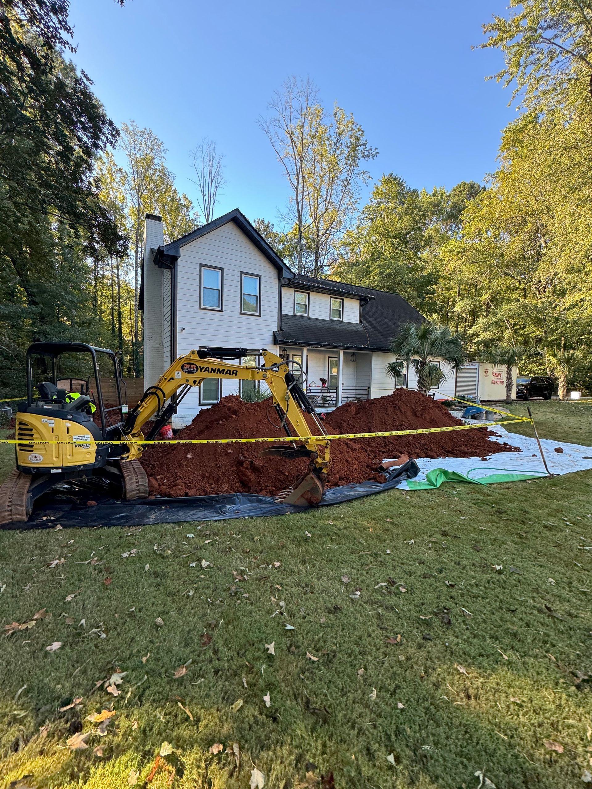 A small excavator next to a house with a pile of reddish-brown material on a tarp.