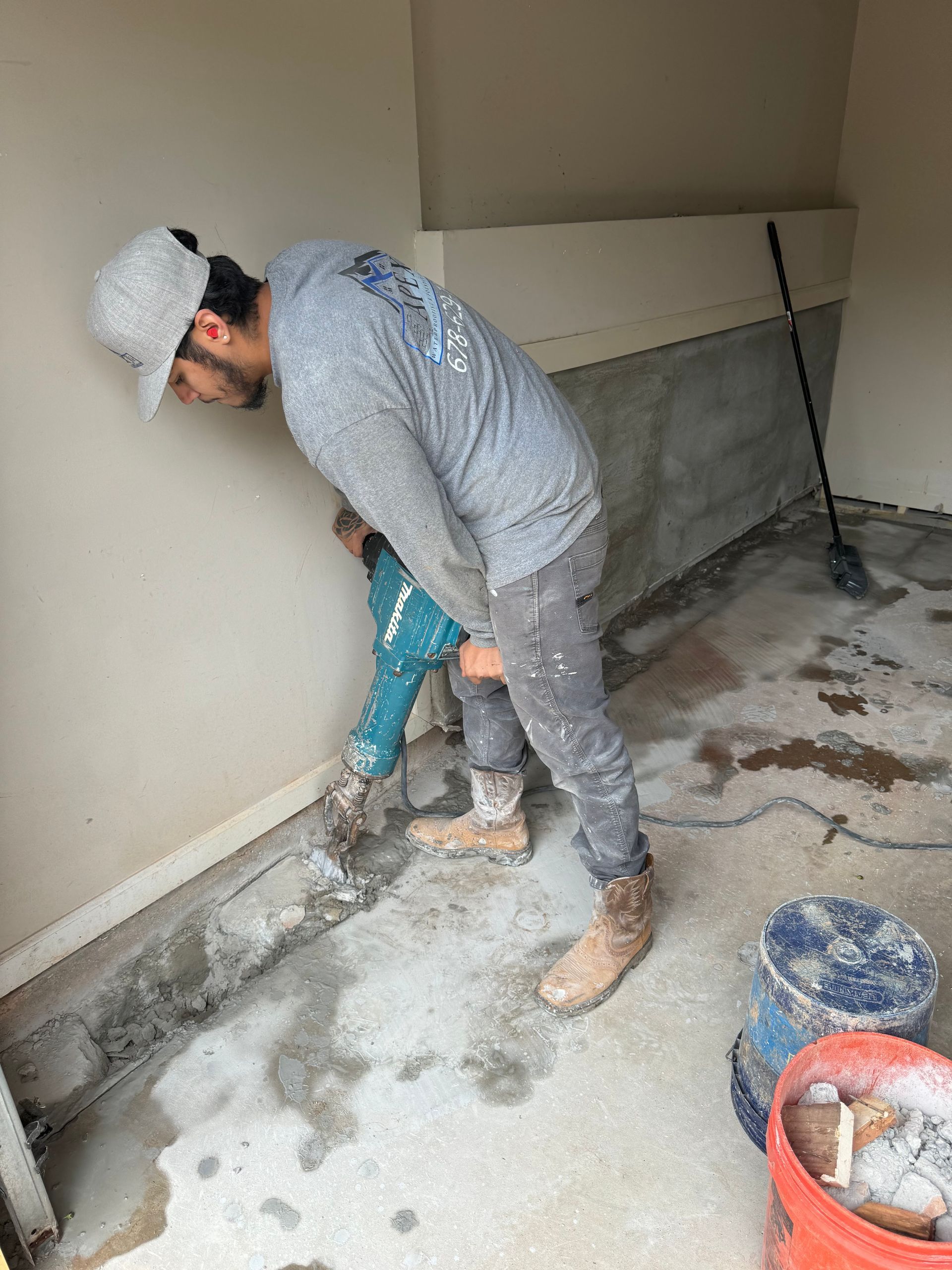 Man using a jackhammer to break up concrete in a room. He wears a hat, long sleeve shirt, and work boots.