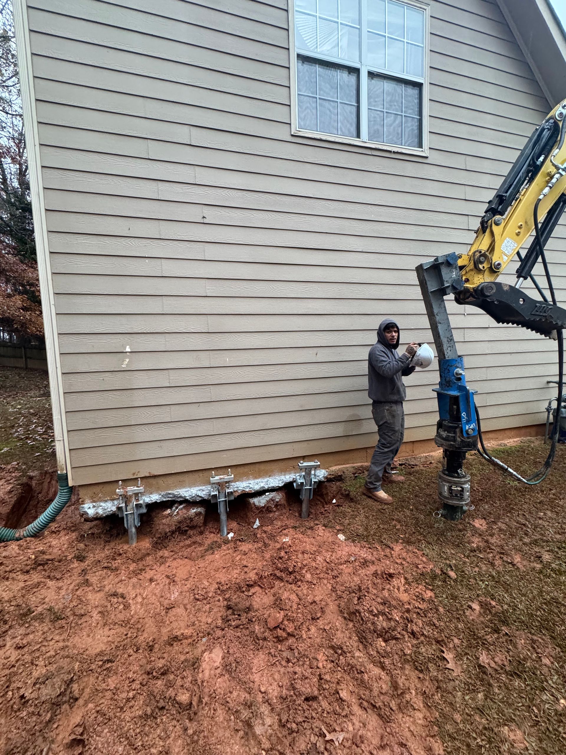 Man operating machinery near a building foundation. Earth is dug up, with a machine attached to the wall.
