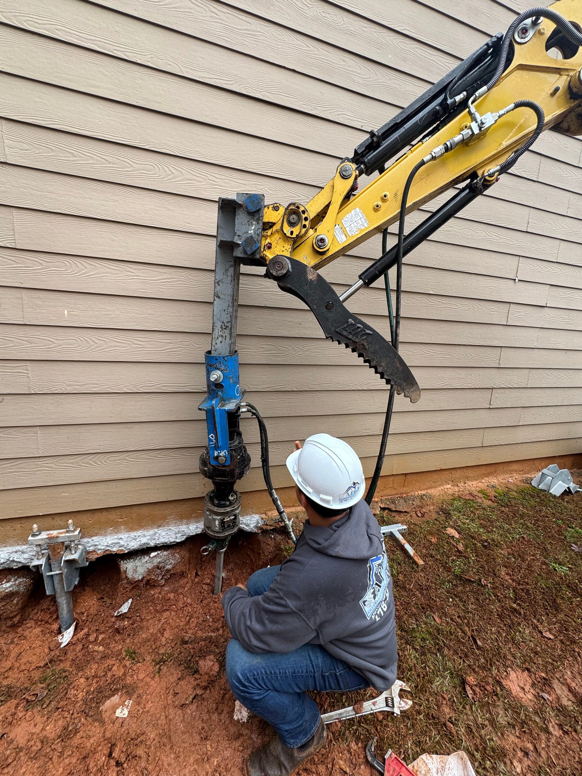 Worker in hard hat operating machinery near a building foundation. Excavator arm extends above.