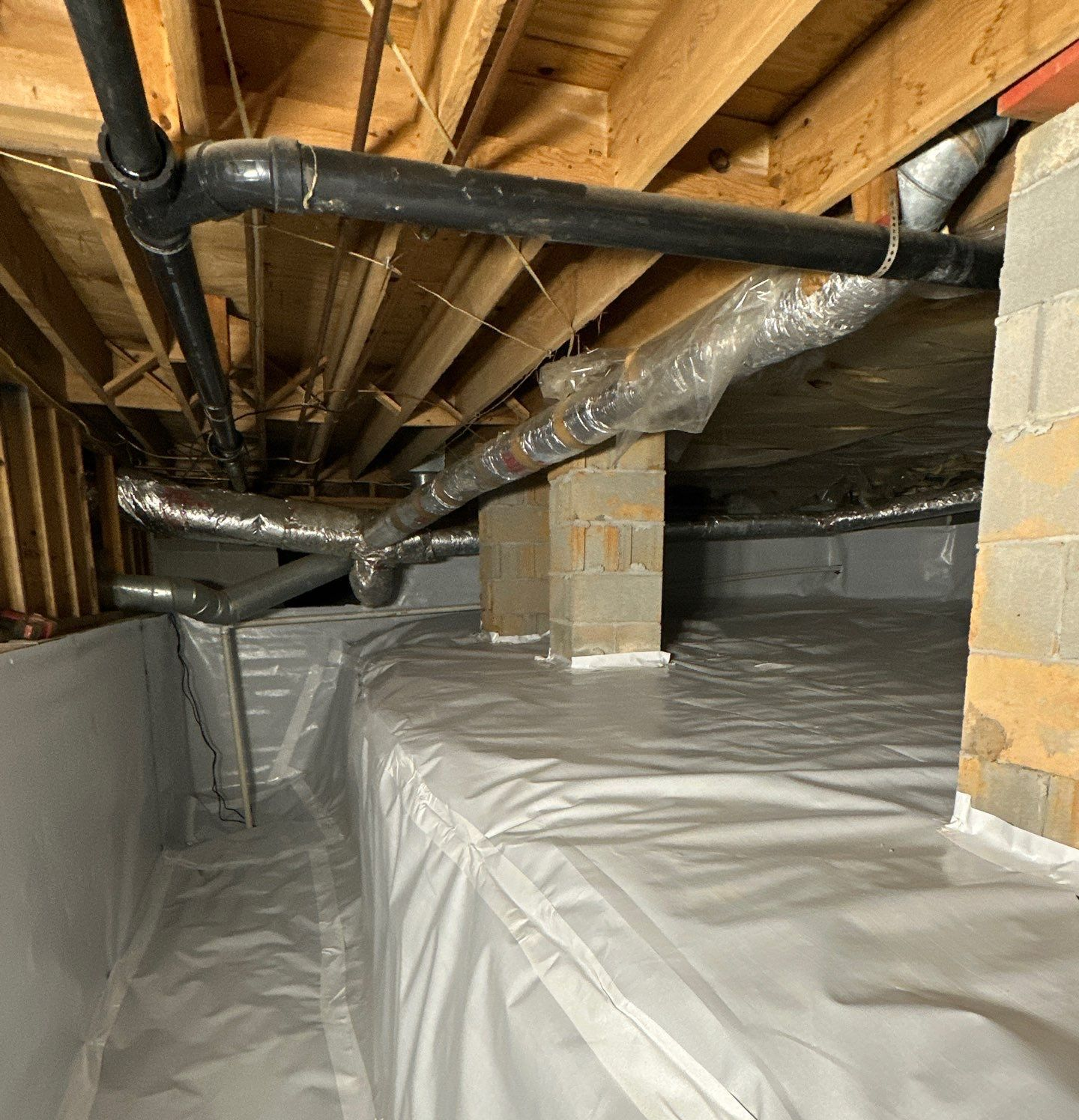 A crawl space featuring a white vapor barrier, exposed wooden joists, black plumbing pipes, and insulated ductwork.