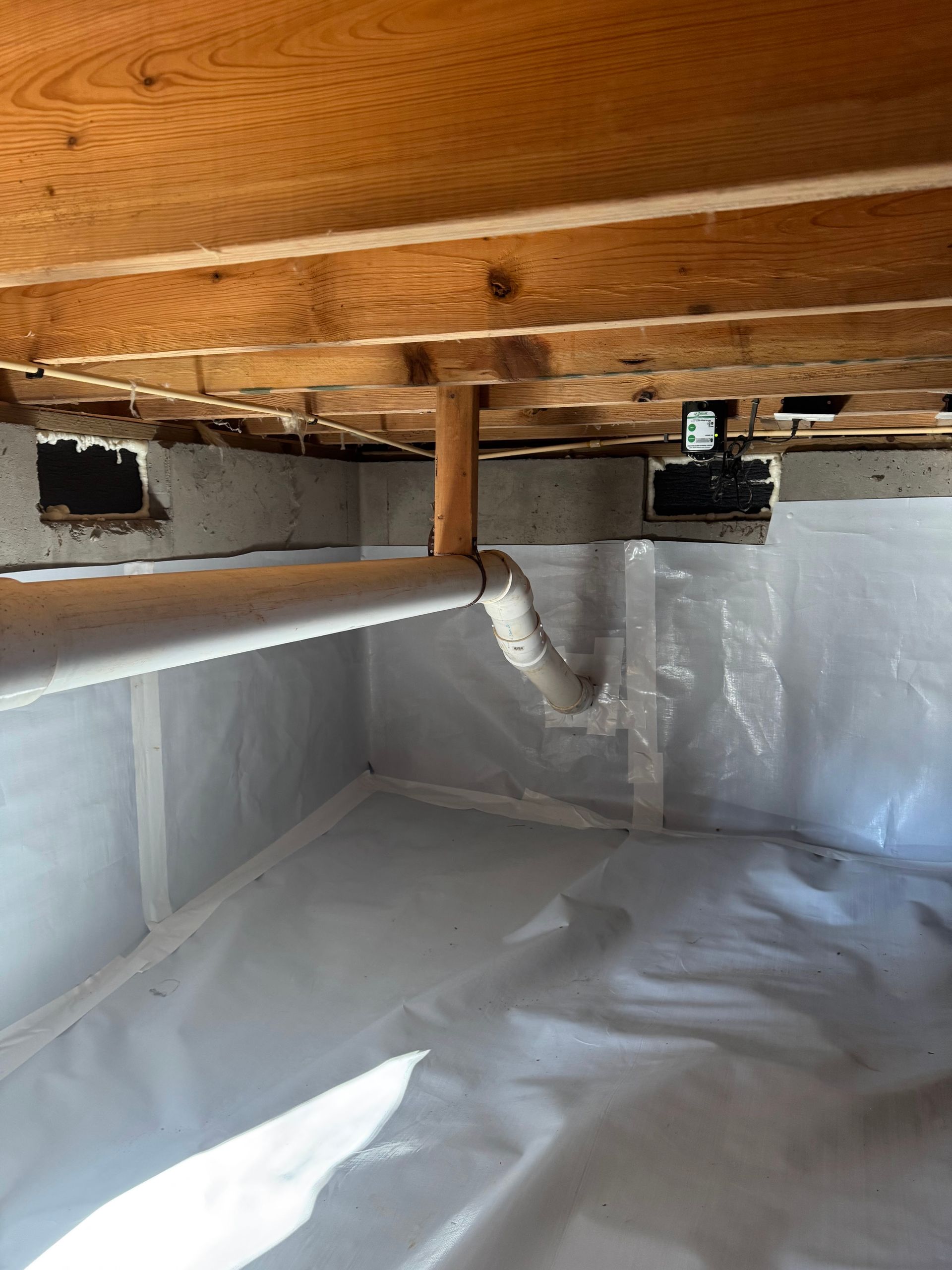 Crawl space with white plastic sheeting on the walls and floor, wooden beams overhead, and plumbing pipes.