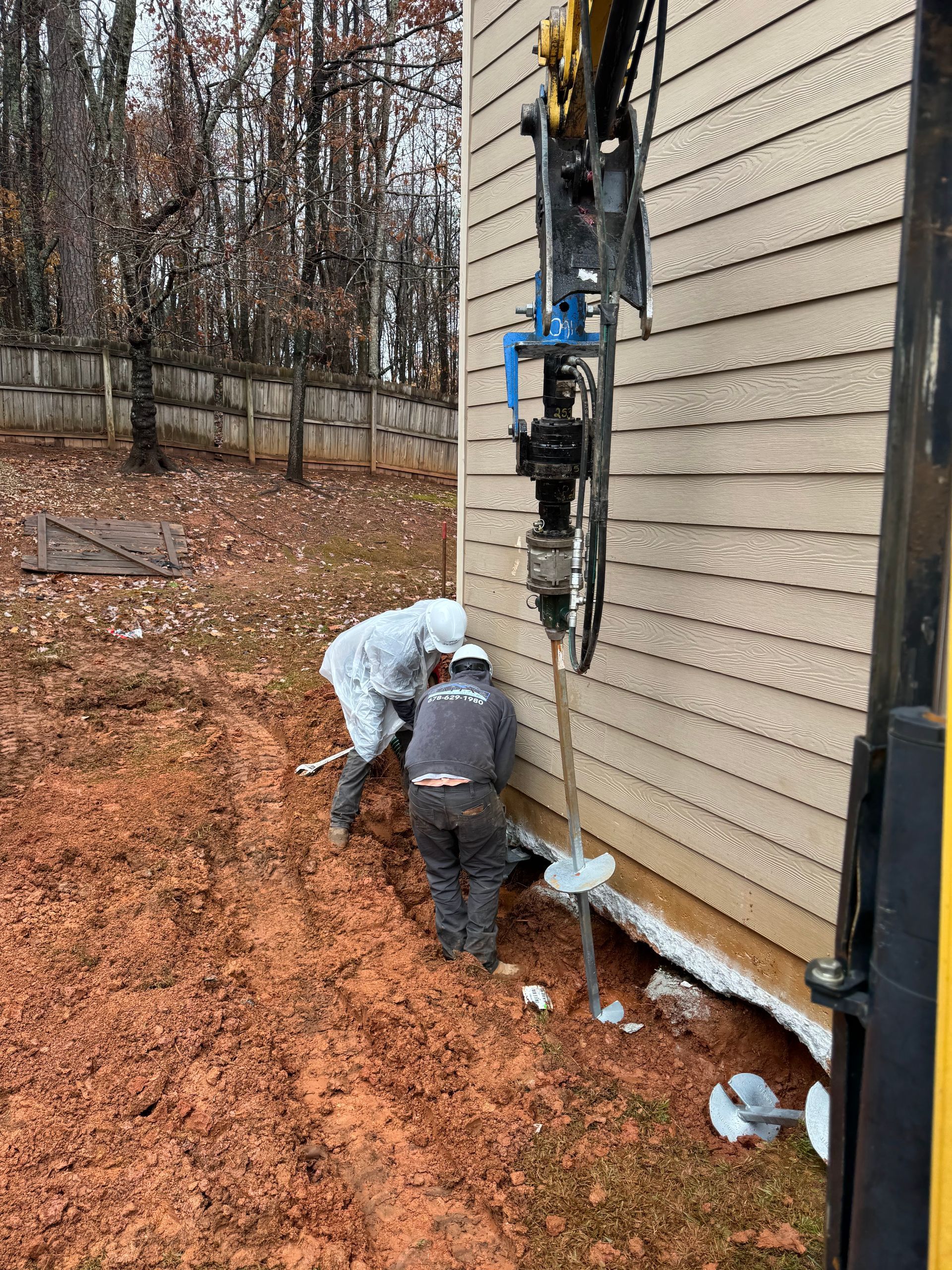 Construction workers using a machine to install a helical pile next to a tan house with brown siding.