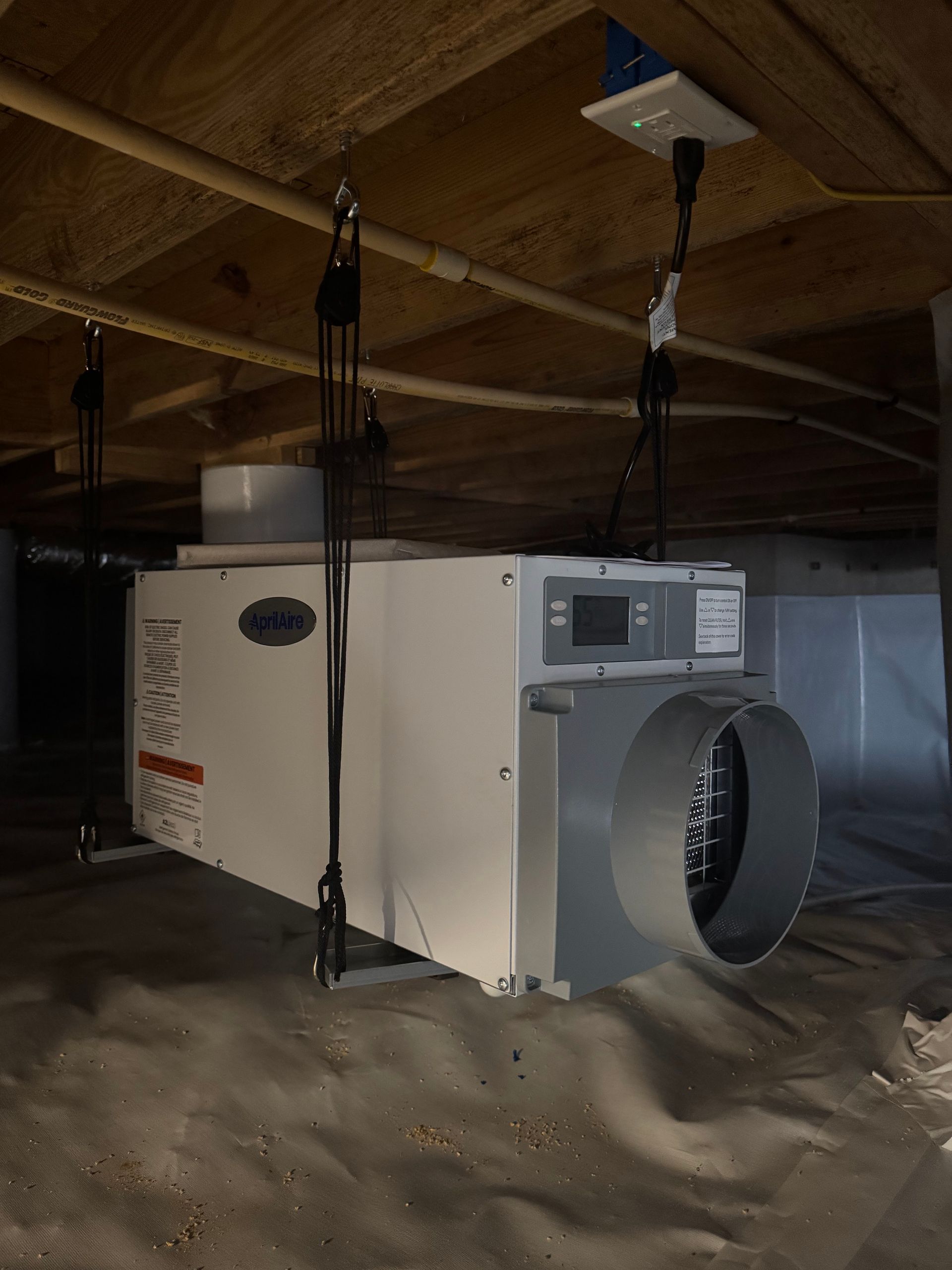 Dehumidifier suspended from joists in crawl space, with ductwork and control panel visible.