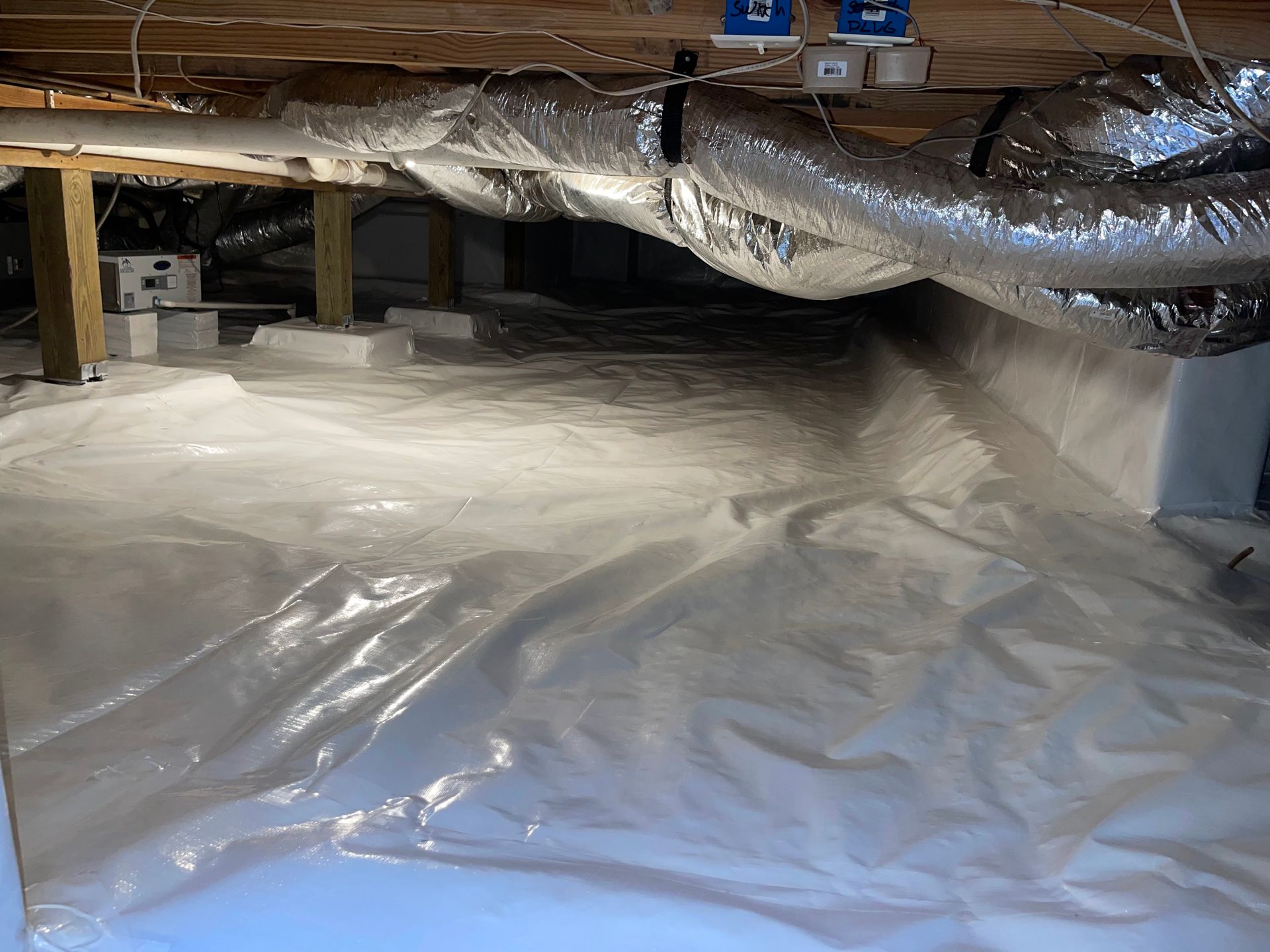 Crawl space with white vapor barrier, wooden beams, HVAC ductwork, and insulation.