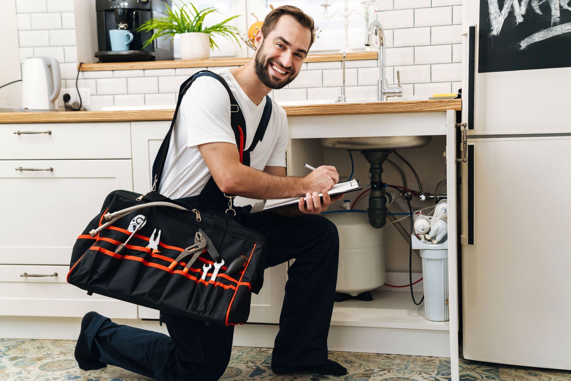 Plumber kneeling in kitchen with tool bag, writing notes near open sink cabinet.