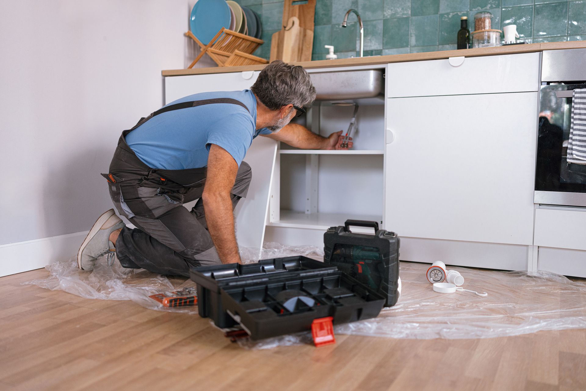 Person kneeling, working inside kitchen cabinet with toolbox open nearby. Person kneeling, working inside kitchen cabinet with toolbox open nearby.
