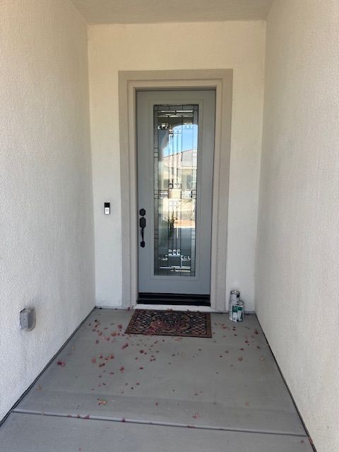 Front door of a home. Gray door with glass panel, recessed entryway, concrete floor, white stucco walls.