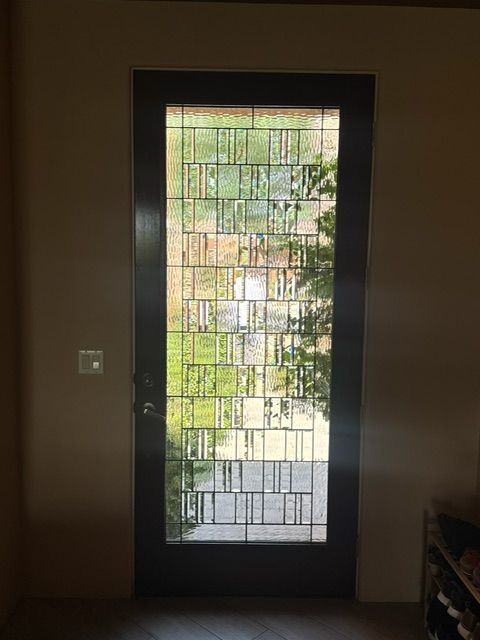 Dark front door with decorative glass pane, beige walls, and sunlight shining through.
