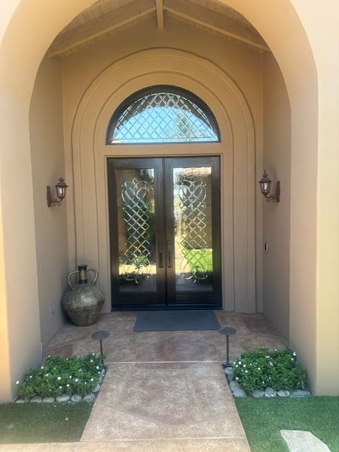 Tan arched doorway with dark double doors and decorative glass, flanked by sconces and landscaping.