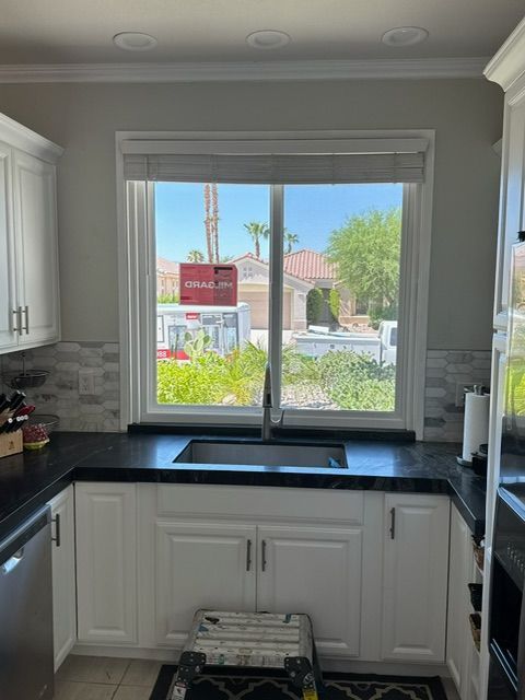 Kitchen with a window overlooking a suburban neighborhood, white cabinets, black countertop, and a partially visible ladder.