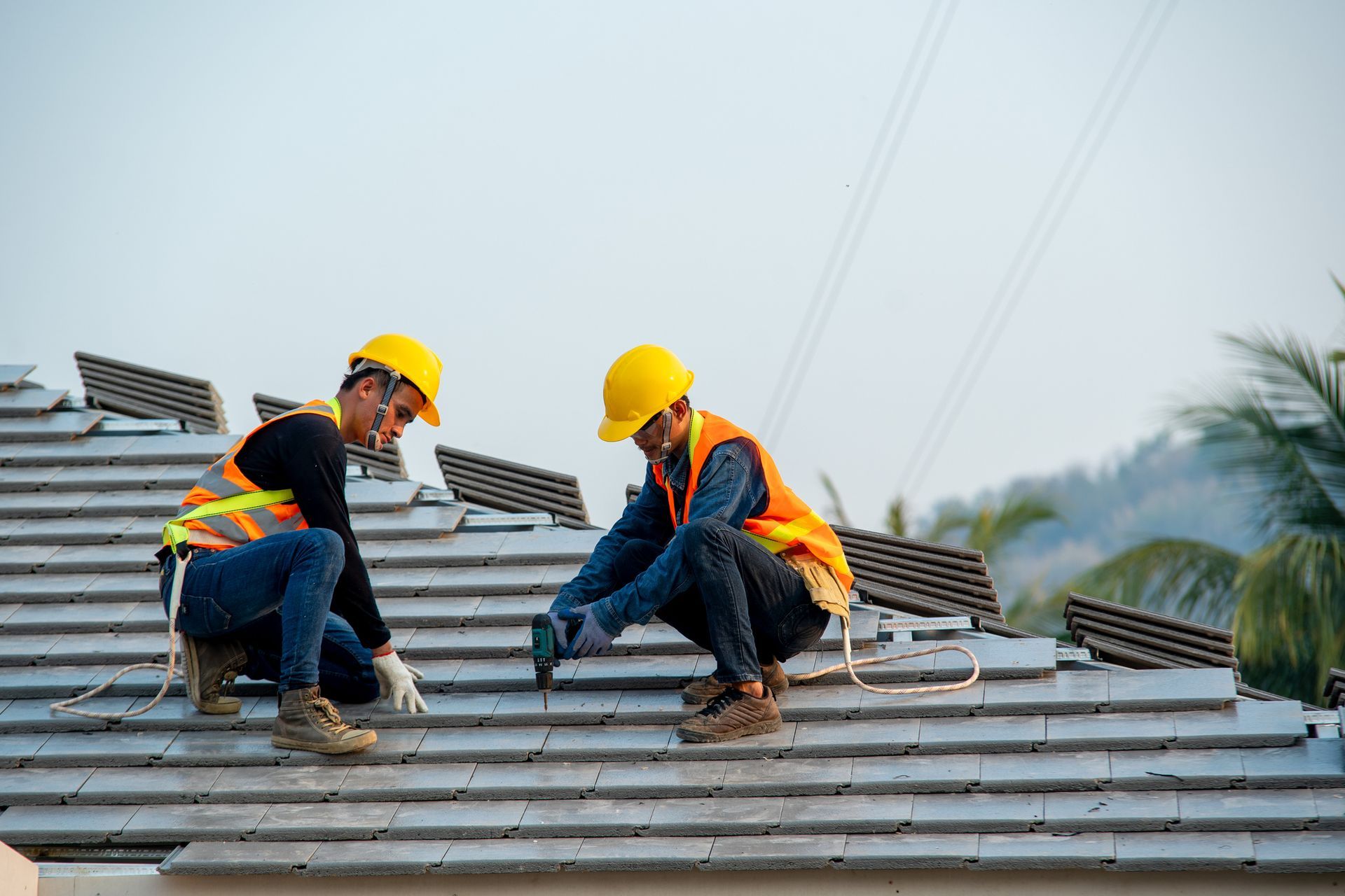 Two construction workers installing roof tiles, wearing hard hats and safety vests.