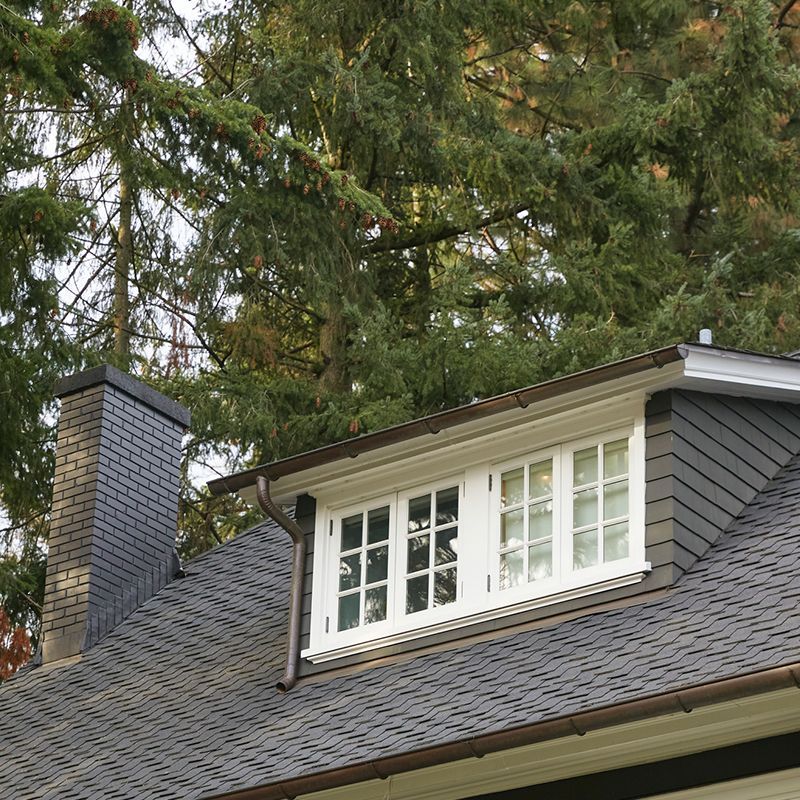 Dormer window on a dark shingled roof with a brick chimney, white trim, and green trees.