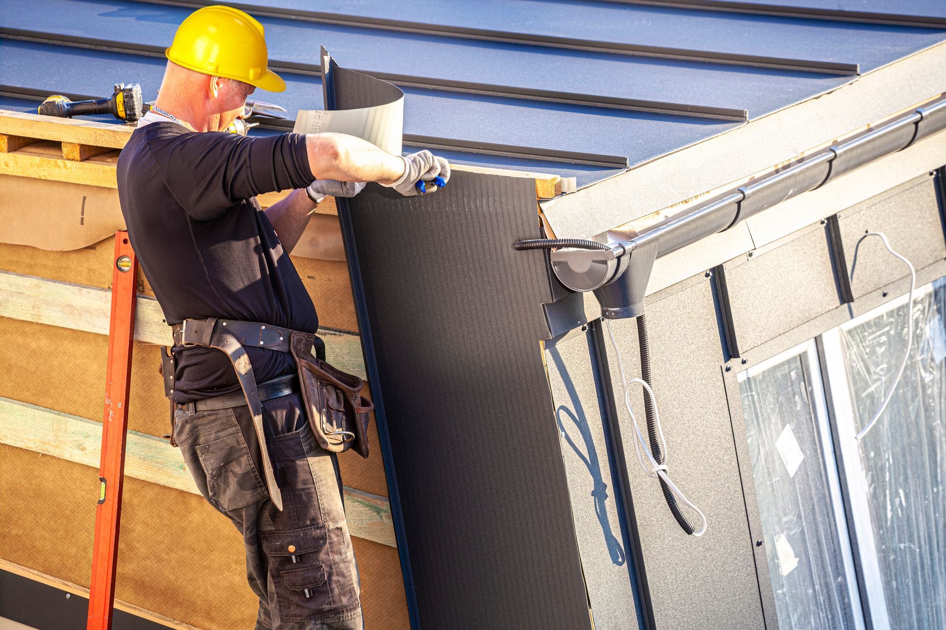 Roofer in yellow hard hat installs black wall panels on a building with a rain gutter, outdoors on a sunny day. Roofer in yellow hard hat installs black wall panels on a building with a rain gutter, outdoors on a sunny day.