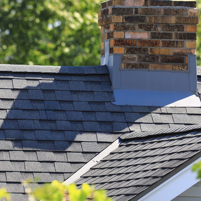 Dark gray shingled roof with brick chimney and flashing against a backdrop of green foliage.