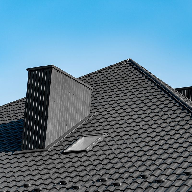 Black tile roof with chimney and skylight against a blue sky.
