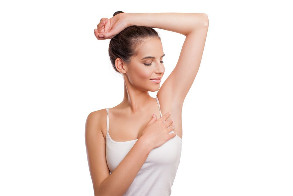 Woman in a white tank top raises arm, touches armpit, smiles. White background.