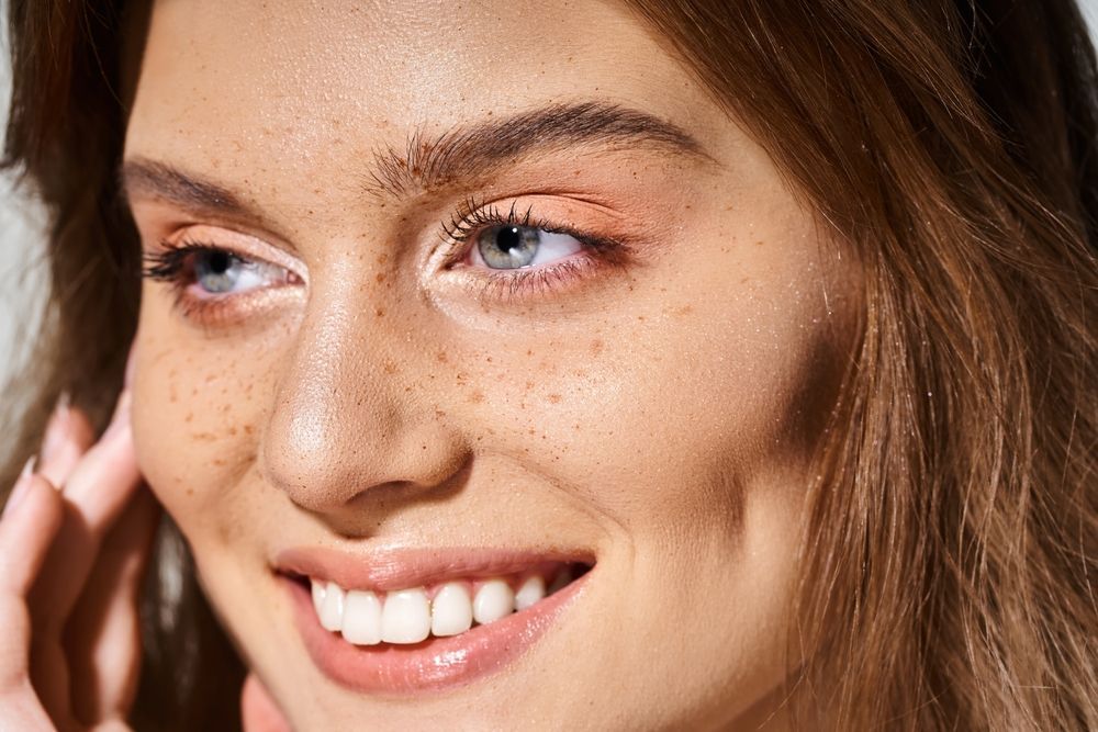 Woman with freckles smiles, looking left. Brown hair, peach eyeshadow, dimple.