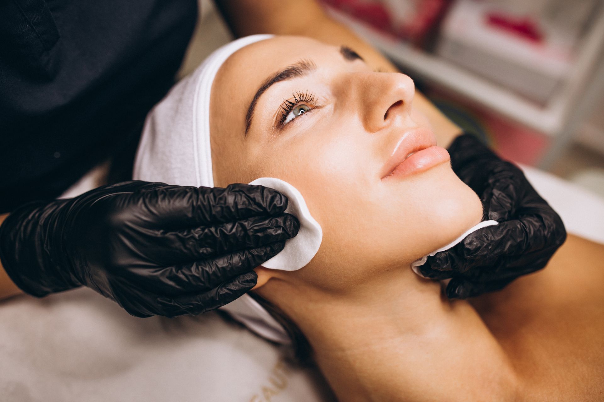 Person receiving facial treatment, with gloved hands cleaning skin with cotton pads.