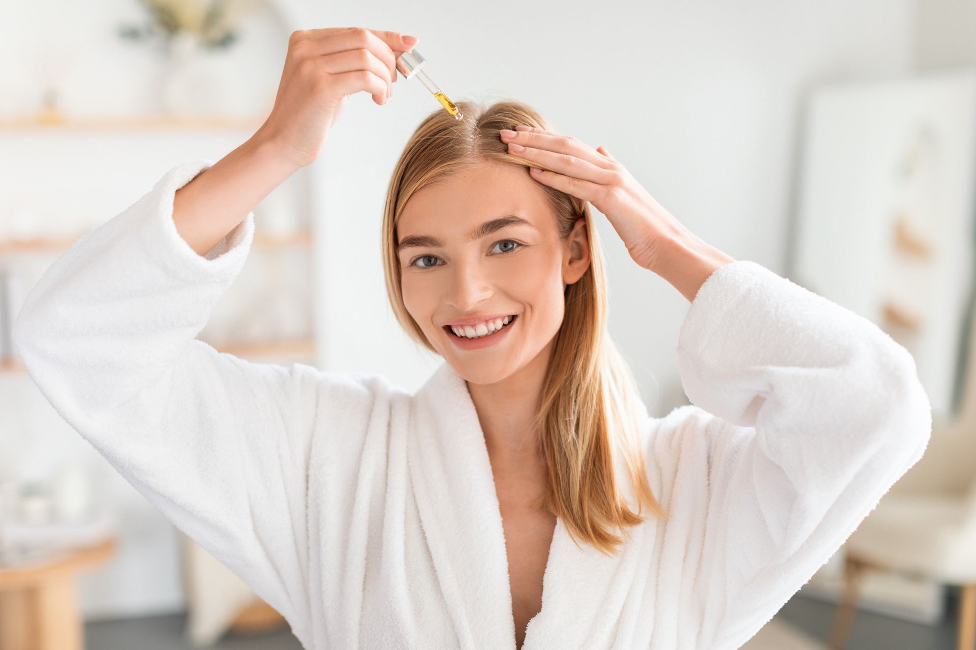 A woman in a bathrobe is applying a serum to her hair.
