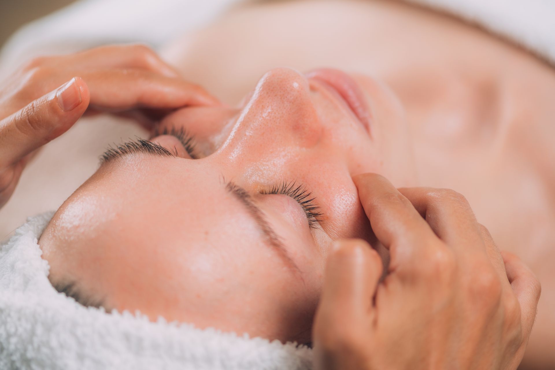 Hands giving a facial massage to a person resting with eyes closed under a white towel